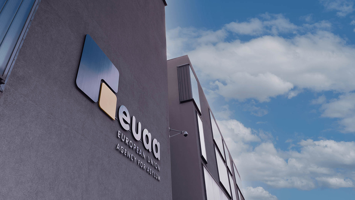 Budova nové Agentury EU pro otázky azylu. Na stěně budovy je vedle textu „EUAA – Agentura Evropské unie pro otázky azylu“ (v angličtině) umístěno logo (velký modrý čtverec, jehož pravou dolní čtvrtinu tvoří vložený žlutý čtverec). V pozadí je vidět modrá obloha.